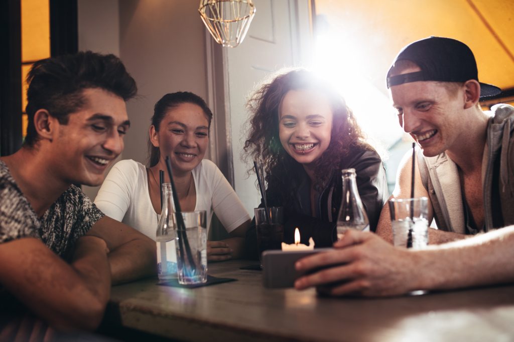 Group of friends sitting together in a cafe looking at mobile phone and smiling. Young guy showing something to his friends on his smart phone.